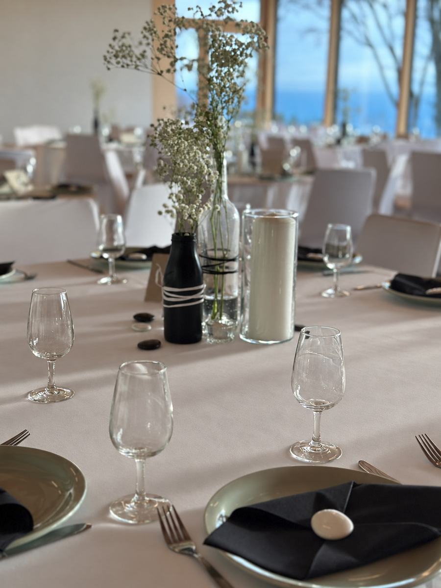 Table de réception dressée devant de grandes baies vitrées : verres, serviette noire, galet blanc et bouquet de gypsophile