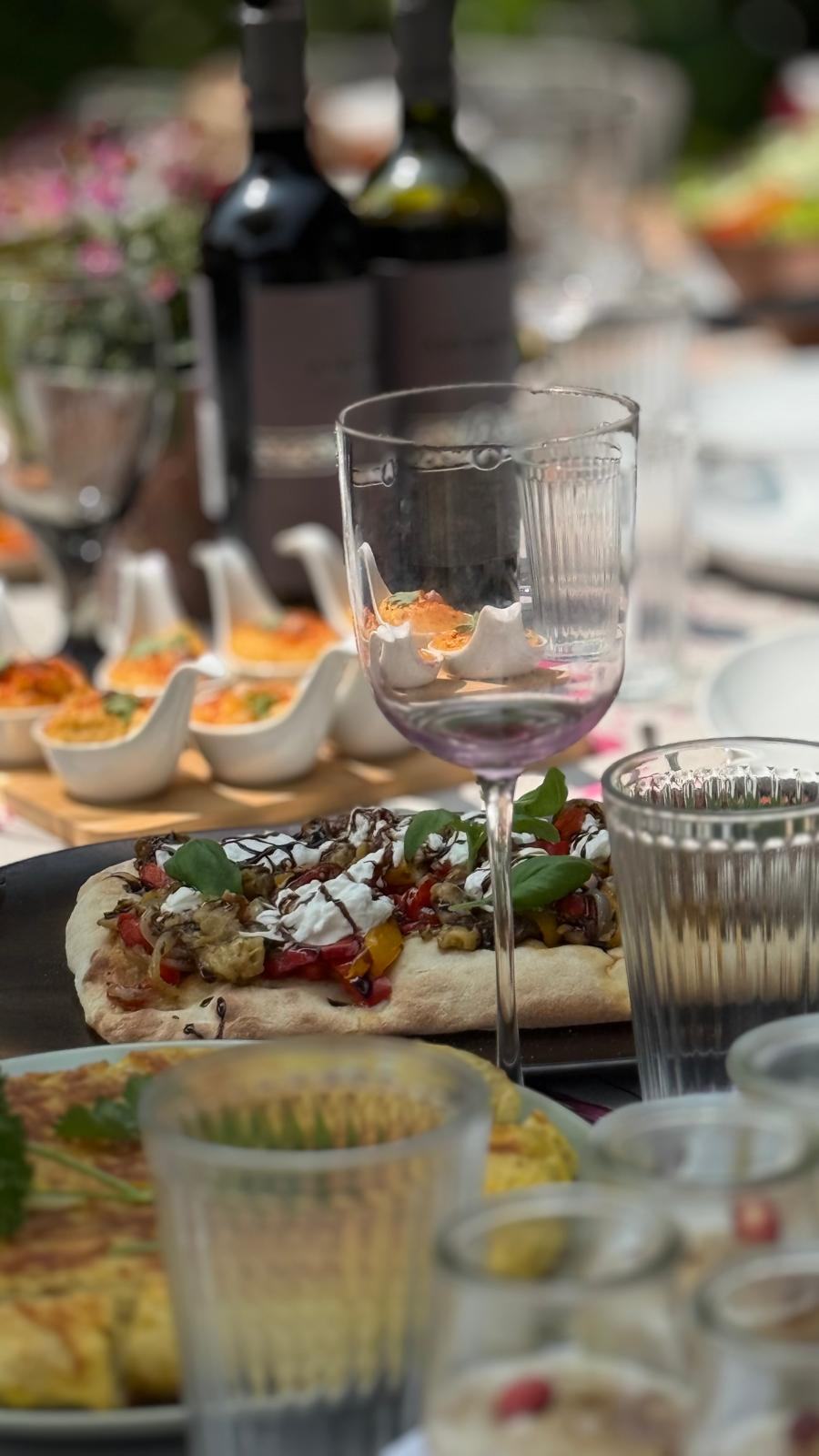 Table de brunch en plein air : focaccia aux légumes grillés et burrata, verrines de houmous au paprika, verres et bouteilles de vin